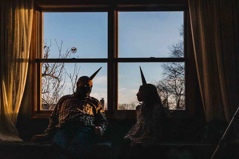 grandfather talking to his granddaughter on her birthday, wearing unicorn party hats and covid masks, sitting in the window partially silhouetted at sunset