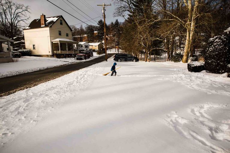 little boy in snowsuit walks through snowy front yard carrying a broom, body language dejected