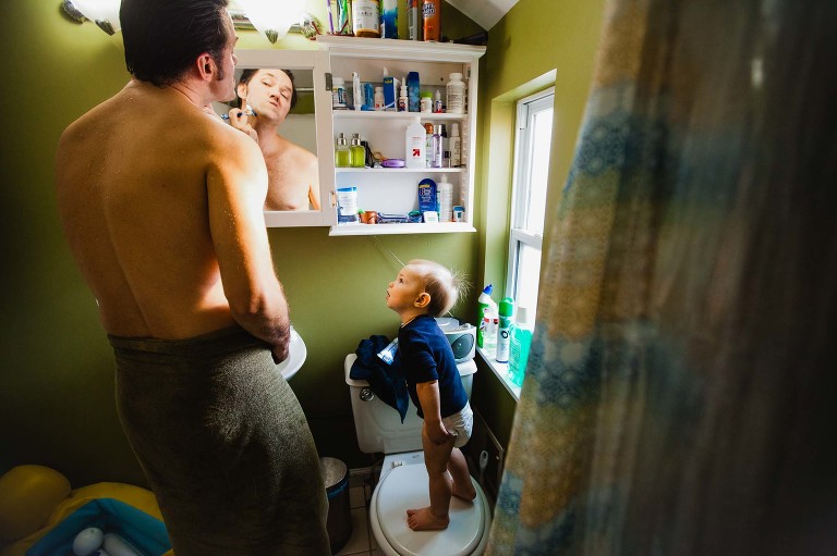dad shaves in the bathroom mirror while his son stands on the toilet staring up at him watching with interest.