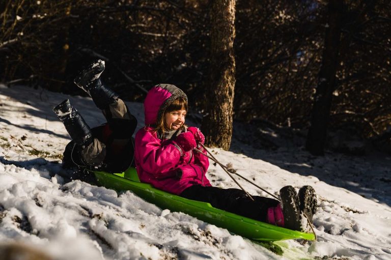 girl laughing and sledding while her friend falls off the sled with his feet in the air.